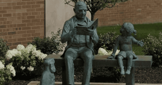 Outdoor statue of an adult reading a book with a child seated nearby, surrounded by flowers outside a library building