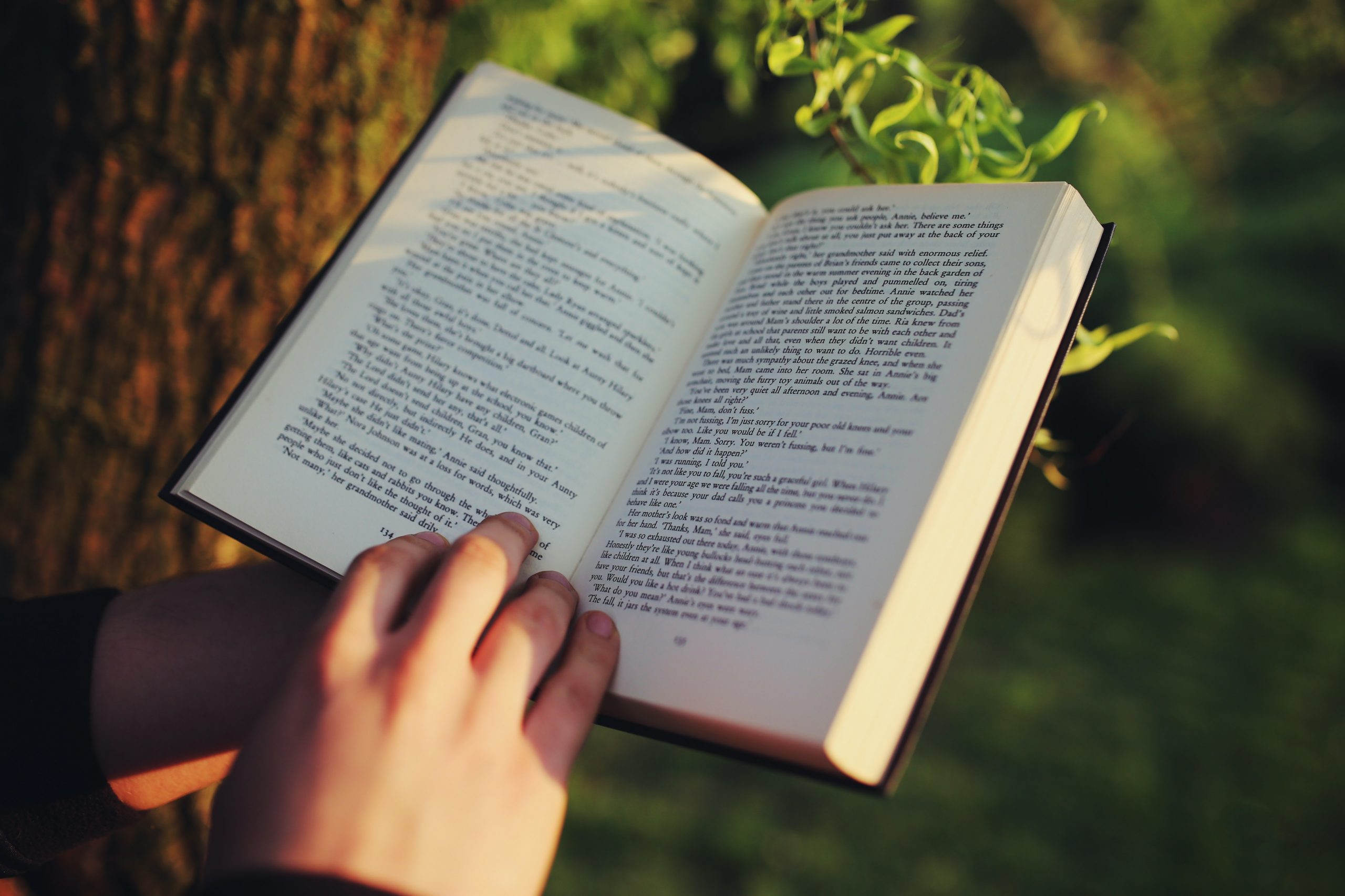 person reading an open book outdoors in sunlight with trees and greenery in the background