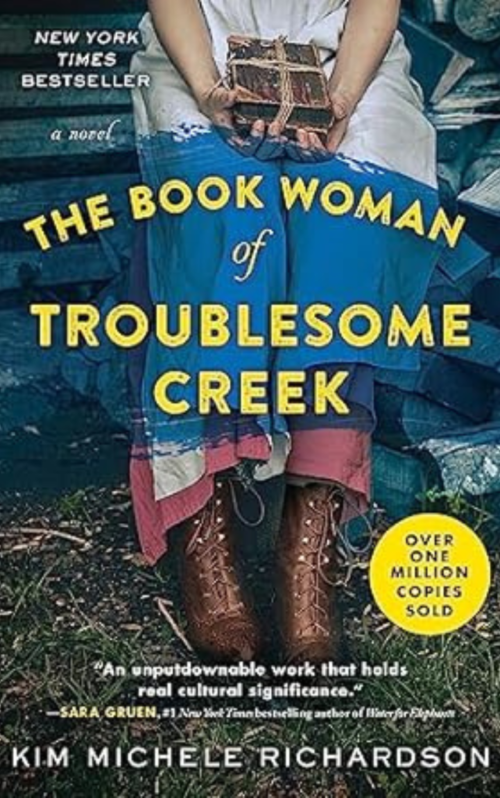Book cover of 'The Book Woman of Troublesome Creek' by Kim Michele Richardson showing a woman holding a small package while seated, with a blue dress and boots visible